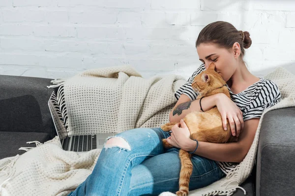 Feliz joven mujer sosteniendo rojo gato mientras utilizando portátil en casa — Stock Photo