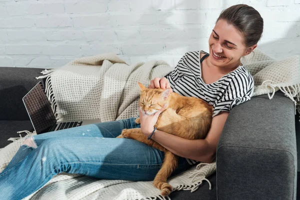 Hermosa feliz joven sentada en sofá y acariciando gato rojo - foto de stock