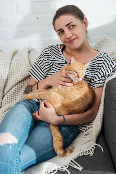 Hermosa sonriente joven mujer sosteniendo rojo gato y sentado en sofá - foto de stock