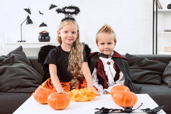Little kids in halloween costumes sitting on sofa at table with pumpkins at home — Stock Photo