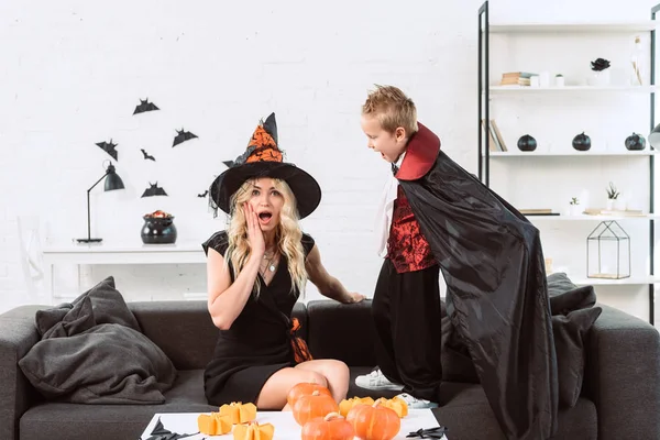 Pequeño niño en traje de vampiro gritándole a la madre en traje de Halloween bruja en casa - foto de stock