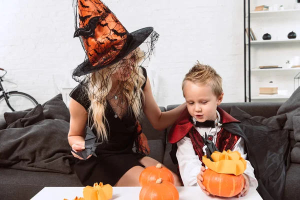 Portrait of mother and son in halloween costumes sitting at coffee table with pumpkins at home — Stock Photo