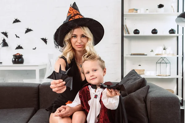 Madre e hijo pequeño en trajes de Halloween mostrando murciélagos de papel negro en el sofá en casa - foto de stock