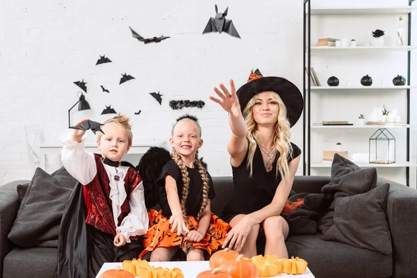 Portrait de mère et d'enfants en costumes d'Halloween jetant des chauves-souris en papier noir à la maison — Photo de stock