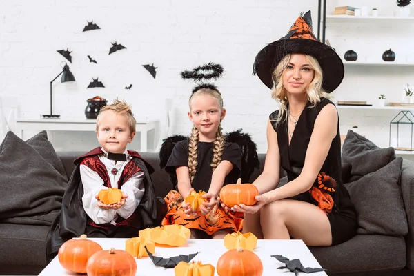 Porträt von Mutter und Kindern in Halloween-Kostümen mit Kürbissen in den Händen, die zu Hause auf dem Sofa sitzen — Stockfoto
