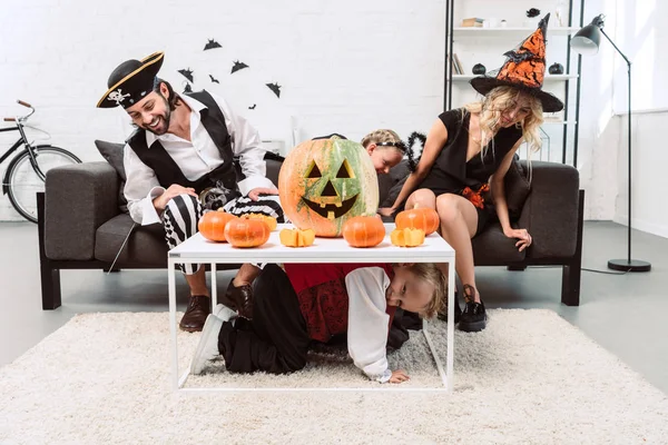 Niño pequeño escondiéndose de los padres y la hermana en trajes de Halloween debajo de la mesa con calabazas en casa - foto de stock