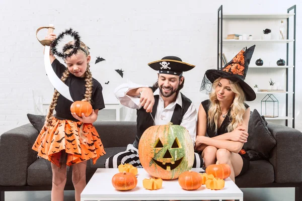 Family in halloween costumes on sofa at coffee table with pumpkins at home — Stock Photo