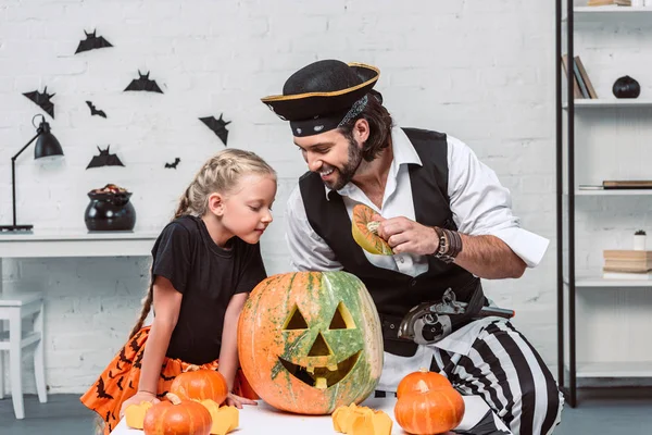 Father and daughter in halloween costumes looking into pumpkin together at home — Stock Photo
