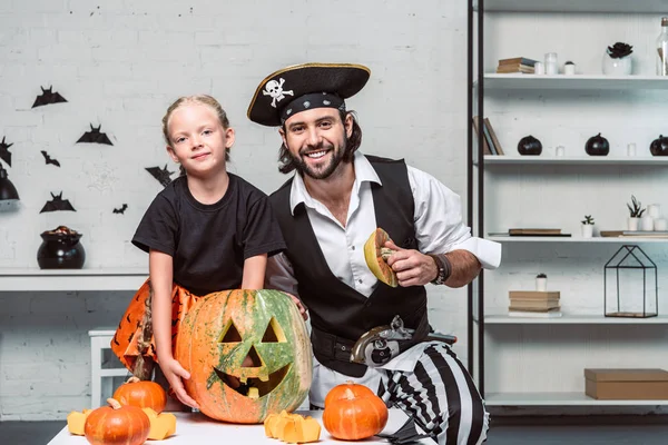 Retrato de padre e hija sonrientes en trajes de Halloween en la mesa con calabaza en casa - foto de stock