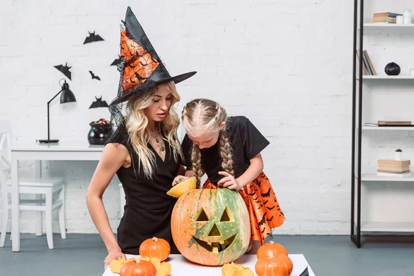 Portrait of mother and little daughter in witches costumes at coffee table with pumpkins at home — Stock Photo