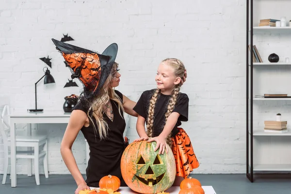 Portrait of mother and little daughter in witches costumes at coffee table with pumpkins at home — Stock Photo