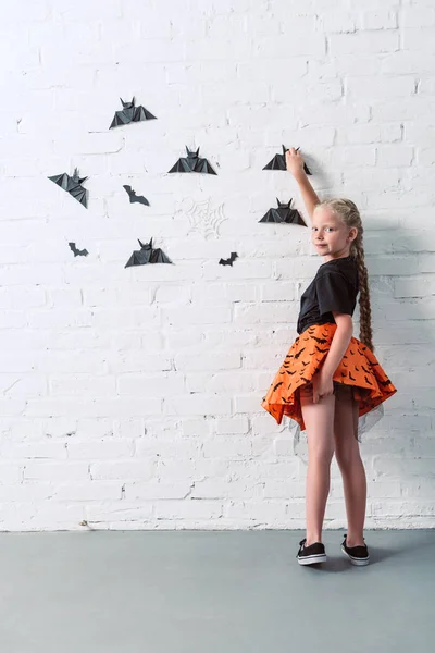 Vista posterior de niño en falda colgando murciélagos de papel negro en la pared de ladrillo blanco, concepto de fiesta de Halloween - foto de stock