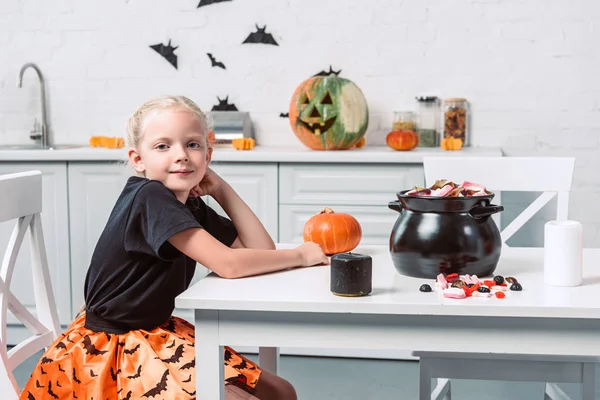Vista lateral do garoto bonito sentado à mesa com guloseimas de Halloween em panela preta em casa — Fotografia de Stock