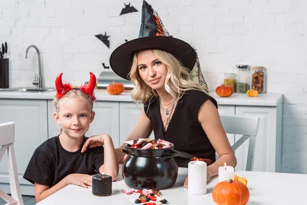 Madre e hija pequeña en trajes de Halloween en la mesa con golosinas en olla negra en la cocina - foto de stock