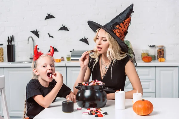 Mutter und kleine Tochter in Halloween-Kostümen essen Leckereien aus schwarzem Topf in der Küche — Stockfoto