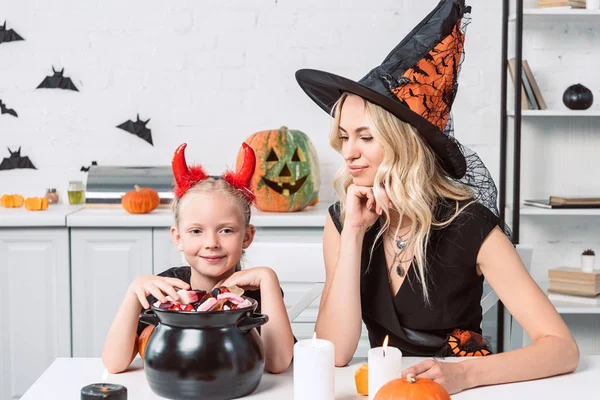 Mother and little daughter in halloween costumes at table with treats in black pot in kitchen — Stock Photo