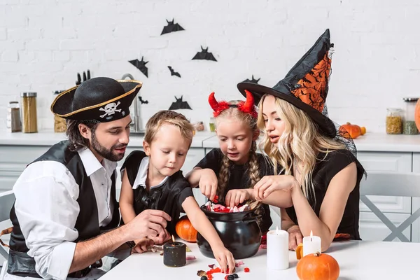 Family in various halloween costumes at table with black pot with treats in kitchen at home — Stock Photo