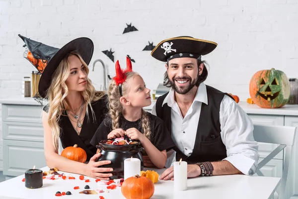 Portrait de parents et fille en costumes d'Halloween à table avec friandises en pot noir dans la cuisine à la maison — Photo de stock