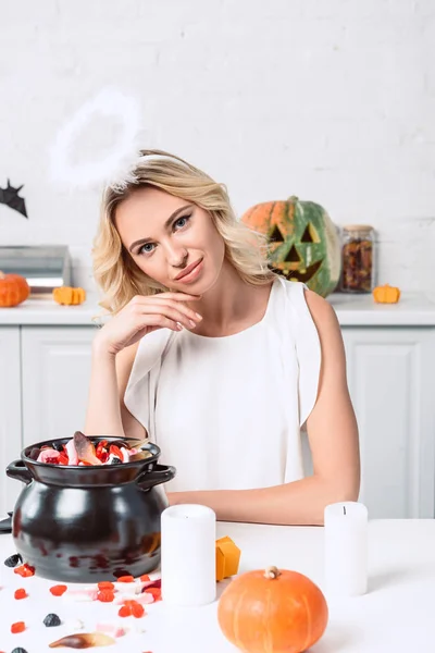 Porträt einer schönen Frau im Engelskostüm, die zu Halloween mit Süßigkeiten im schwarzen Topf am Tisch sitzt — Stockfoto