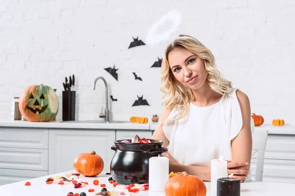 Retrato de mujer hermosa en traje de ángel sentado a la mesa con dulces en olla negra para halloween - foto de stock