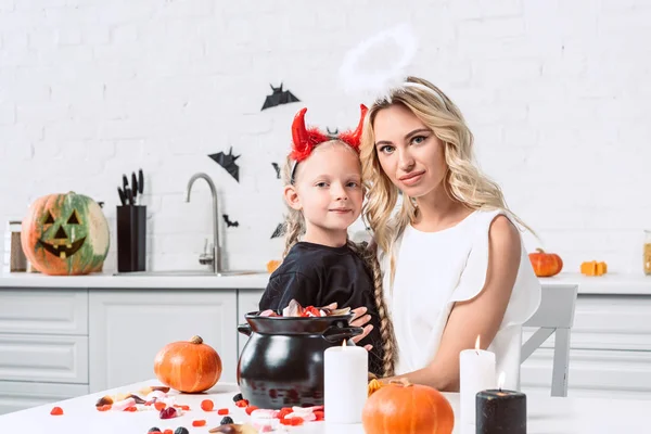 Retrato de madre e hija en trajes de halloween en la mesa con dulces en olla negra en la cocina en casa - foto de stock