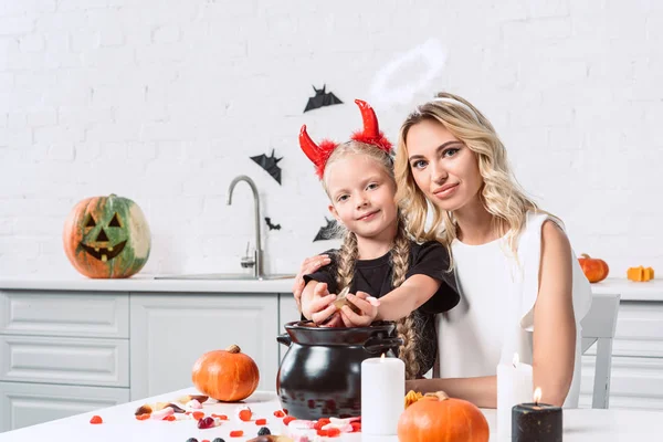 Retrato de madre e hija en trajes de halloween en la mesa con dulces en olla negra en la cocina en casa - foto de stock
