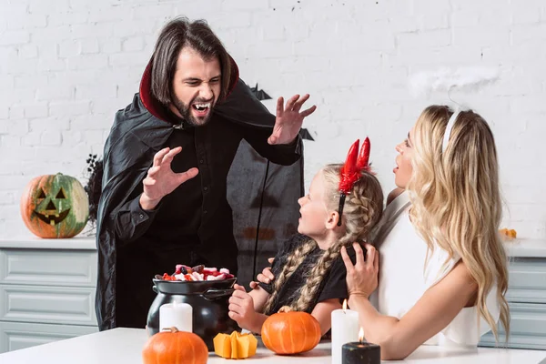 Retrato de los padres y la hija en trajes de Halloween en la mesa con golosinas en olla negra en la cocina en casa - foto de stock