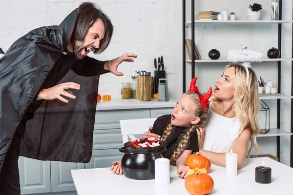 Ritratto di genitori e figlia in costumi di Halloween a tavola con prelibatezze in pentola nera in cucina a casa — Foto stock