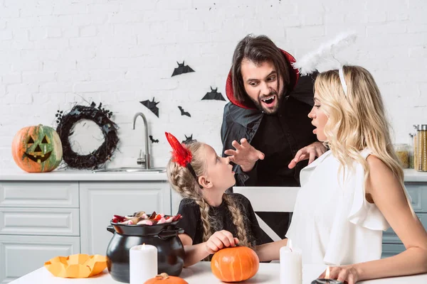 Ritratto di genitori e figlia in costumi di Halloween a tavola con prelibatezze in pentola nera in cucina a casa — Foto stock