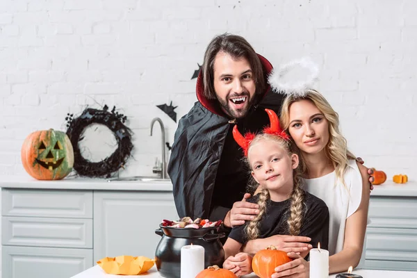 Retrato de los padres y la hija en trajes de Halloween en la mesa con golosinas en olla negra en la cocina en casa - foto de stock