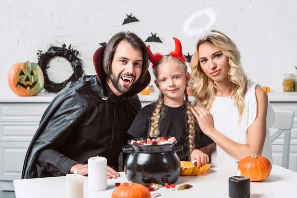 Portrait of parents and daughter in halloween costumes at table with treats in black pot in kitchen at home — Stock Photo