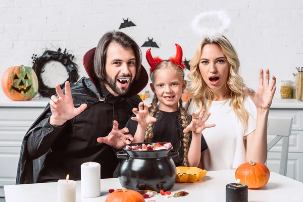 Retrato de los padres y la hija en trajes de Halloween en la mesa con golosinas en olla negra en la cocina en casa - foto de stock