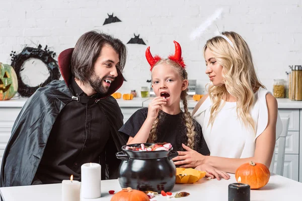 Portrait of parents and daughter in halloween costumes at table with treats in black pot in kitchen at home — Stock Photo