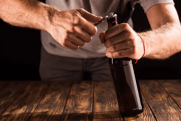Visão parcial do homem que abre a garrafa de cerveja pelo abridor na mesa de madeira — Fotografia de Stock