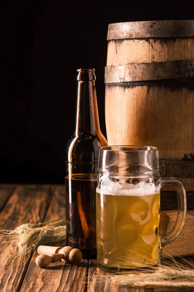 Glass of light beer with foam, beer bottle, wheat and wooden barrel at table on black background — Stock Photo