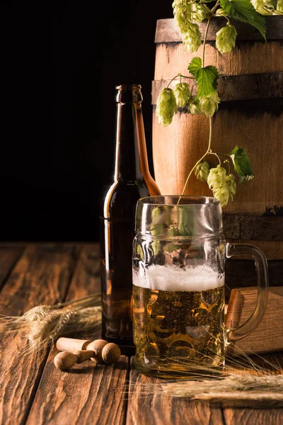 Пивная бутылка, стакан свежего пива с пеной, хмель, пшеница и деревянная бочка за столом на черном фоне — стоковое фото