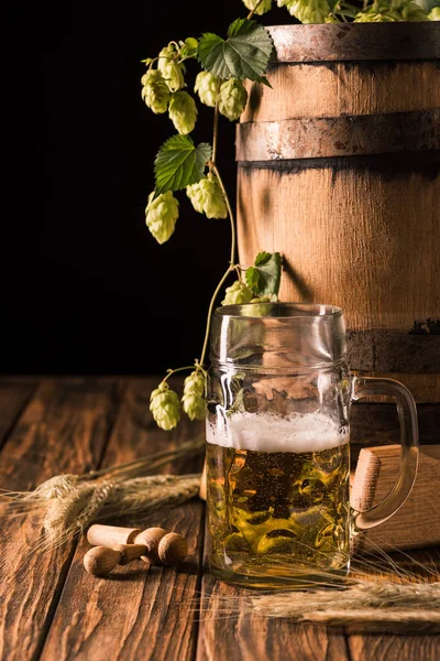 Tasse de bière fraîche avec mousse, blé, houblon et tonneau en bois à table sur fond noir — Photo de stock