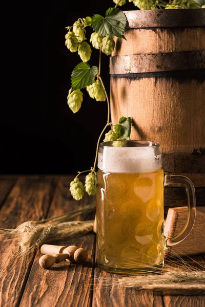 Glas mit frischem Bier, Hopfen, Weizen und Holzfass am Tisch auf schwarzem Hintergrund — Stockfoto