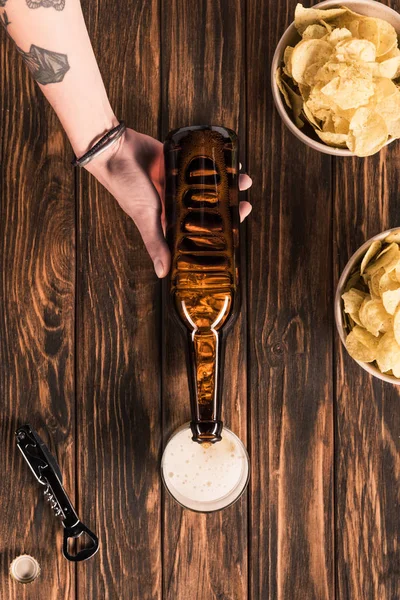 Imagem cortada de mulher derramando cerveja em vidro à mesa de madeira com batatas fritas — Fotografia de Stock