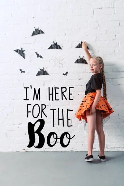 Vista posterior de niño en falda colgando murciélagos de papel negro en la pared de ladrillo blanco, concepto de fiesta de Halloween con letras 