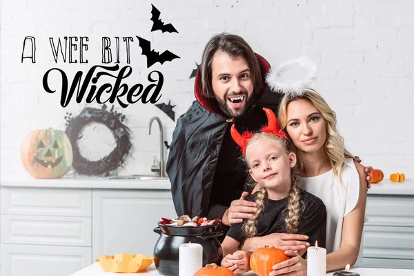 Retrato de los padres y la hija en trajes de Halloween en la mesa con golosinas en olla negra en la cocina en casa con letras 