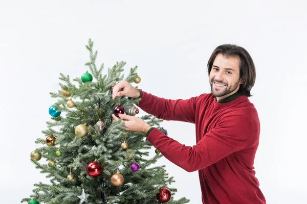Улыбающийся мужчина украшает рождественскую елку стеклянными шариками и смотрит на камеру, изолированную на белом — стоковое фото