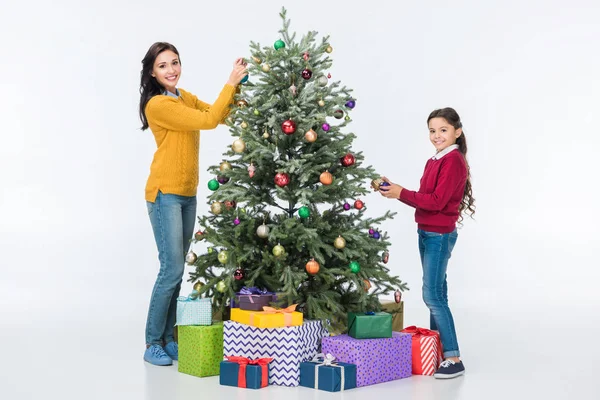 Madre feliz con hija decorando el árbol de Navidad con bolas de vidrio y mirando a la cámara aislada en blanco - foto de stock