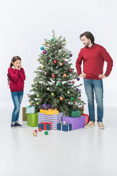 Hija sorprendida mirando en bolas de vidrio mientras el padre de pie cerca del árbol de Navidad con regalos aislados en blanco - foto de stock