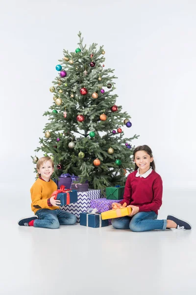 Hermanos sentados cerca del árbol de navidad con regalos y mirando a la cámara aislada en blanco - foto de stock