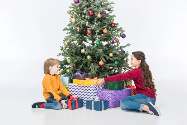 Hermanos sonrientes sentados cerca del árbol de Navidad con regalos aislados en blanco - foto de stock