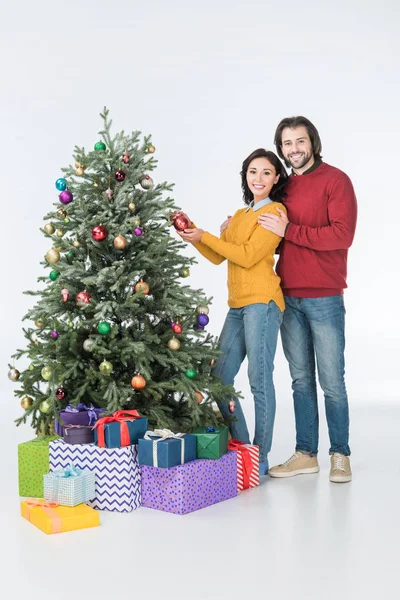 Feliz pareja decorando el árbol de Navidad con regalos y mirando a la cámara aislada en blanco - foto de stock