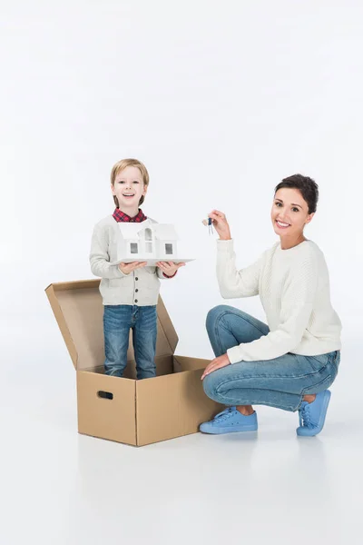 Menino sorrindo segurando casa de papelão com chave mãe segurando de casa nova pronta para mover-se para casa nova isolada no branco — Fotografia de Stock