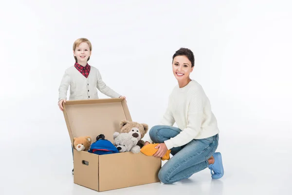 Mère souriante avec son fils assis près d'une boîte en carton avec des jouets et regardant la caméra isolée sur blanc — Photo de stock
