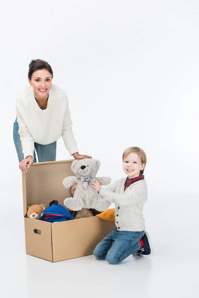 Glückliche Mutter mit Sohn hält Teddybär und sitzt in der Nähe von Karton mit Spielzeug isoliert auf weiß — Stockfoto